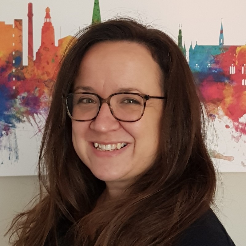 Headshot of Abi, a white woman with long, brown, wavy hair, smiling at the camera. She is wearing a black top and brown tortoiseshell glasses. A colourful painting behind Abi shows landmarks along the Dundee skyline.