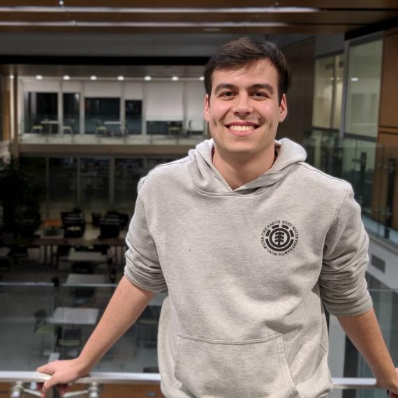 Portrait of Connor Geddes, a white man with short dark hair. He is wearing a grey hoodie and is smiling directly at the camera.