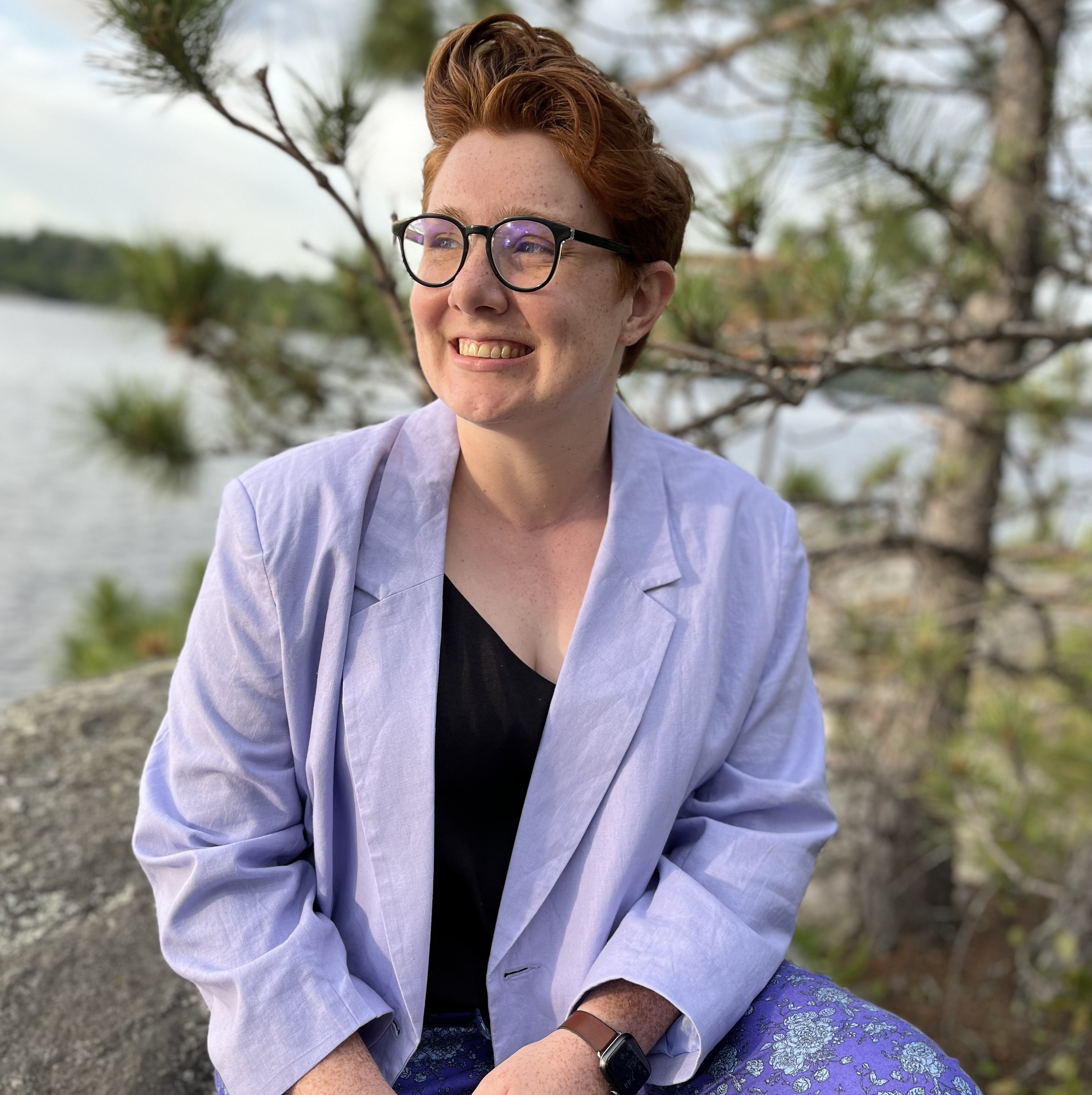 Emma, a white woman with short red hair and black round glasses wearing a purple blazer, black shirt, and purple patterned pants, sits on a rock smiling but not looking at the camera. A tree and water are behind her.