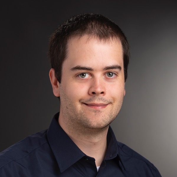 A portrait of Garreth Tigwell, a white male with dark brown hair, smiling and wearing a dark blue shirt, while in front of a dark background.