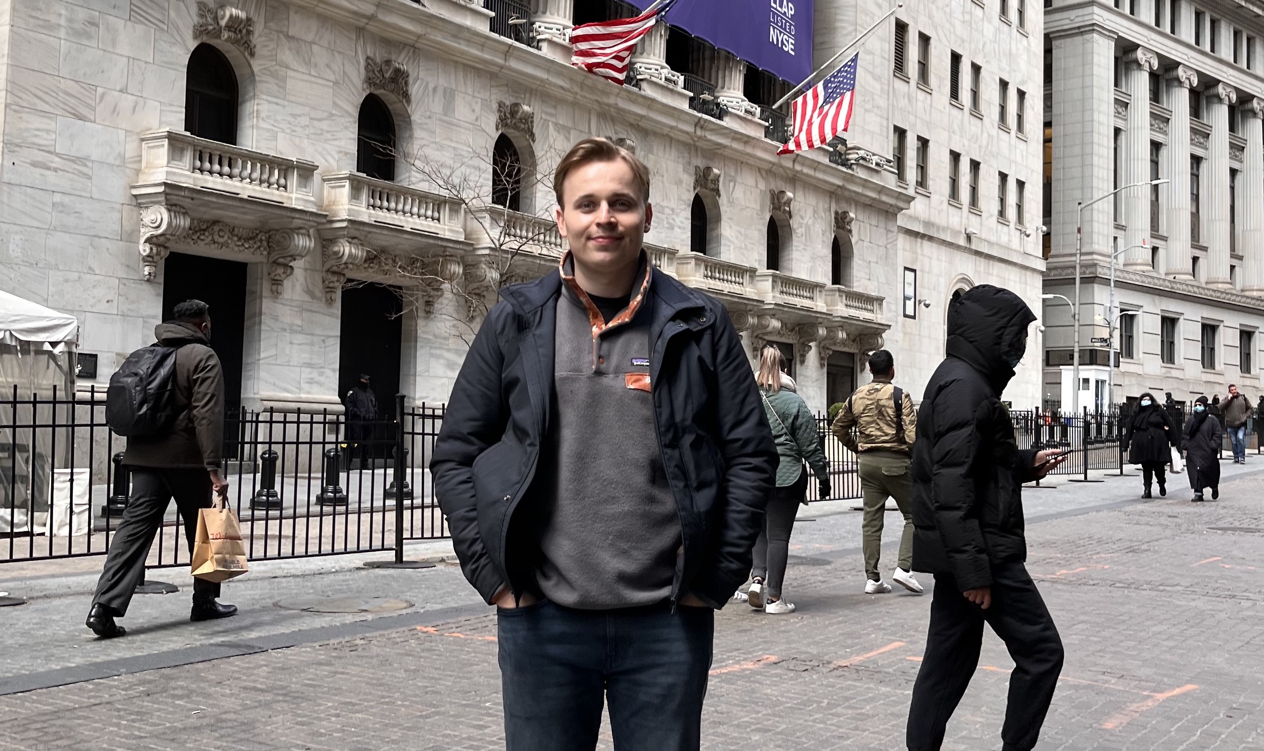 A photograph of Humphrey Curtis, a white male with dark blonde hair, smiling and wearing a grey jumper with dark navy jacket, while in front of a grey and white stone building.