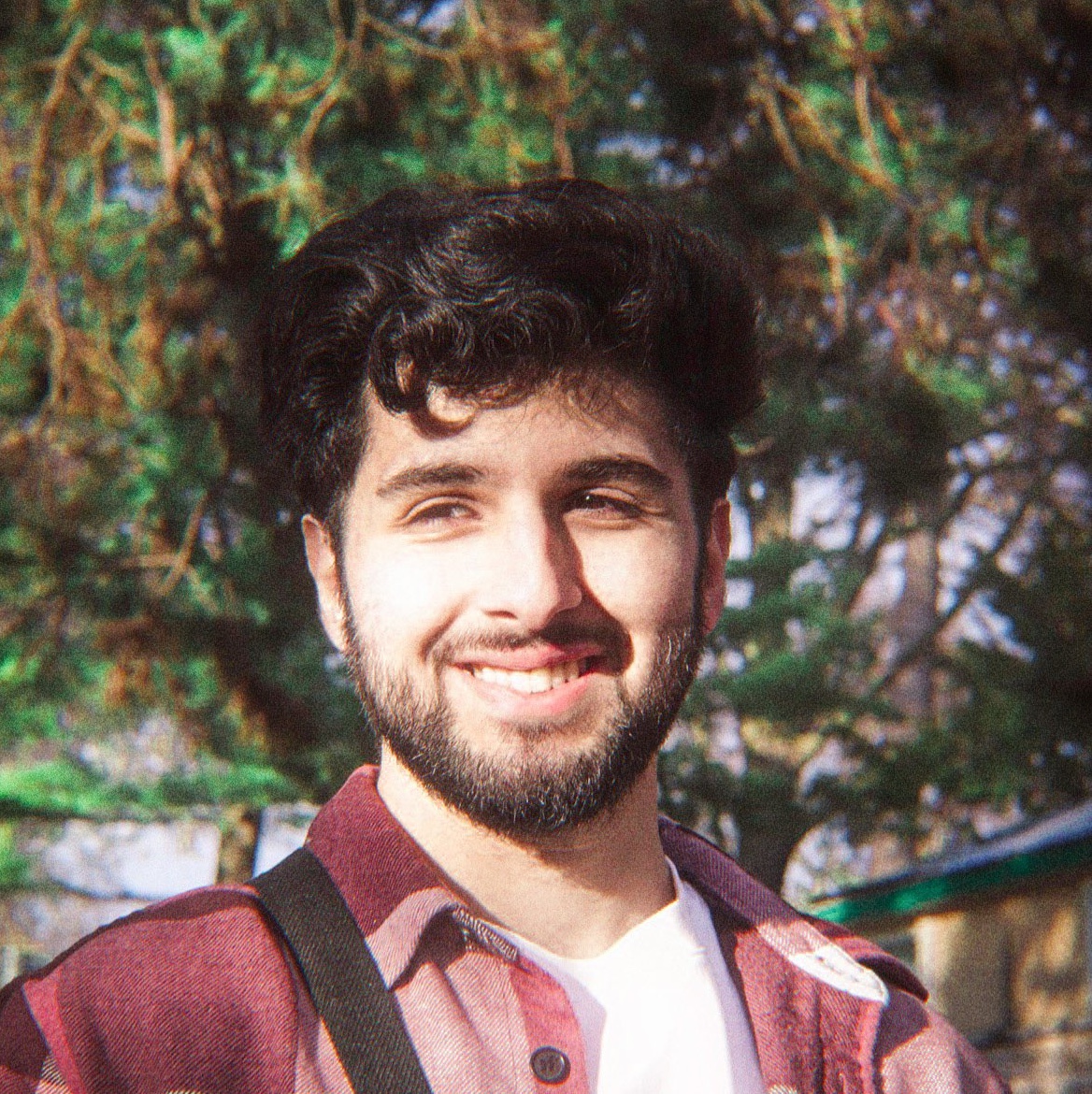 Portrait of Jaipreet Singh, a brown man with curly dark hair and a beard. He is wearing a burgundy flannel over a white t-shirt and is smiling directly at the camera.