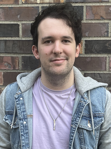 Jesse Martinez, a fair-skinned masc-presenting person with short, wavy brown hair, mild stubble, and a silver eyebrow piercing. He is wearing a pink t-shirt under a denim jacket-hoodie combo and a twisted silver pendant necklace.