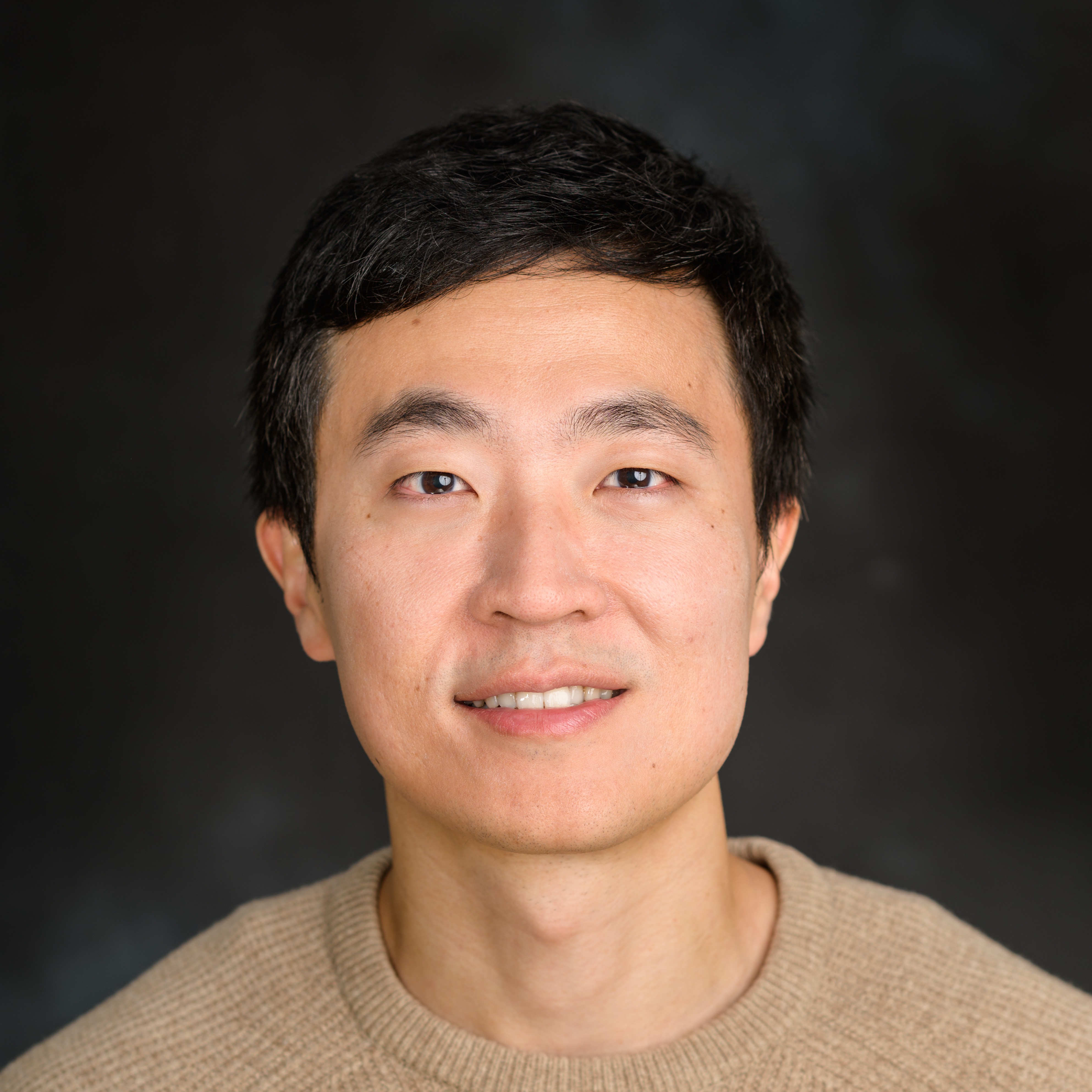 Portrait of a smiling Asian man wearing a beige sweater against a dark background.