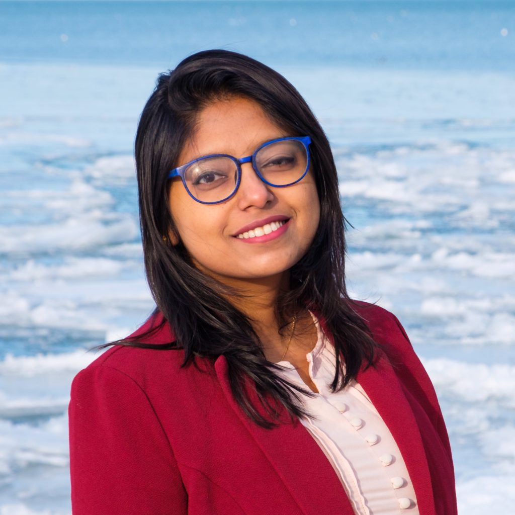 Maitraye, a Brown woman with black shoulder-length straight hair, is smiling at the camera. She is wearing round glasses and a red blazer over a pink shirt.