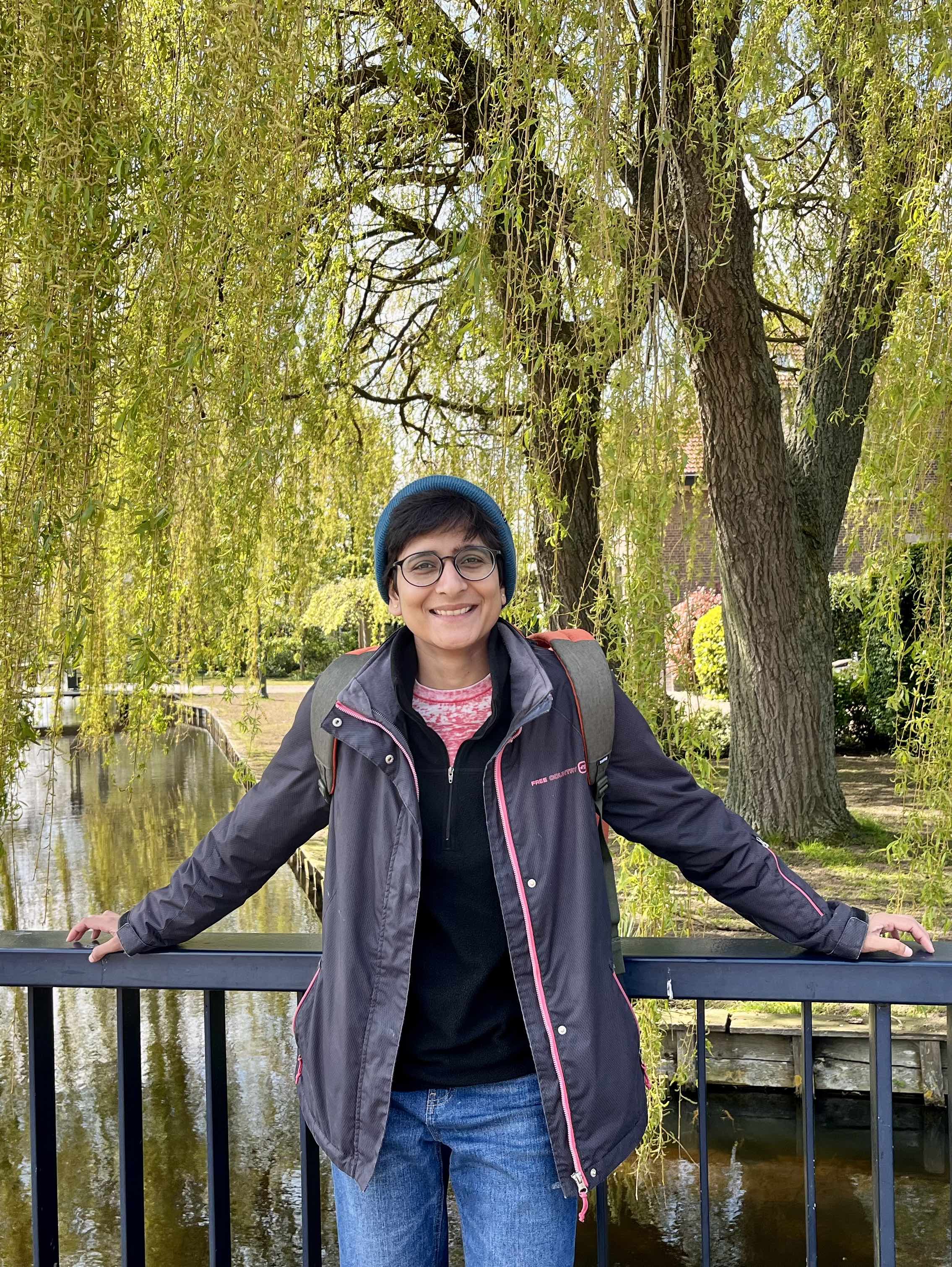 Mauli Pandey, a brown woman, smiling into the camera and standing against a backdrop of green foliage. She is wearing round black glasses, a blue beanie, and a dark grey jacket.