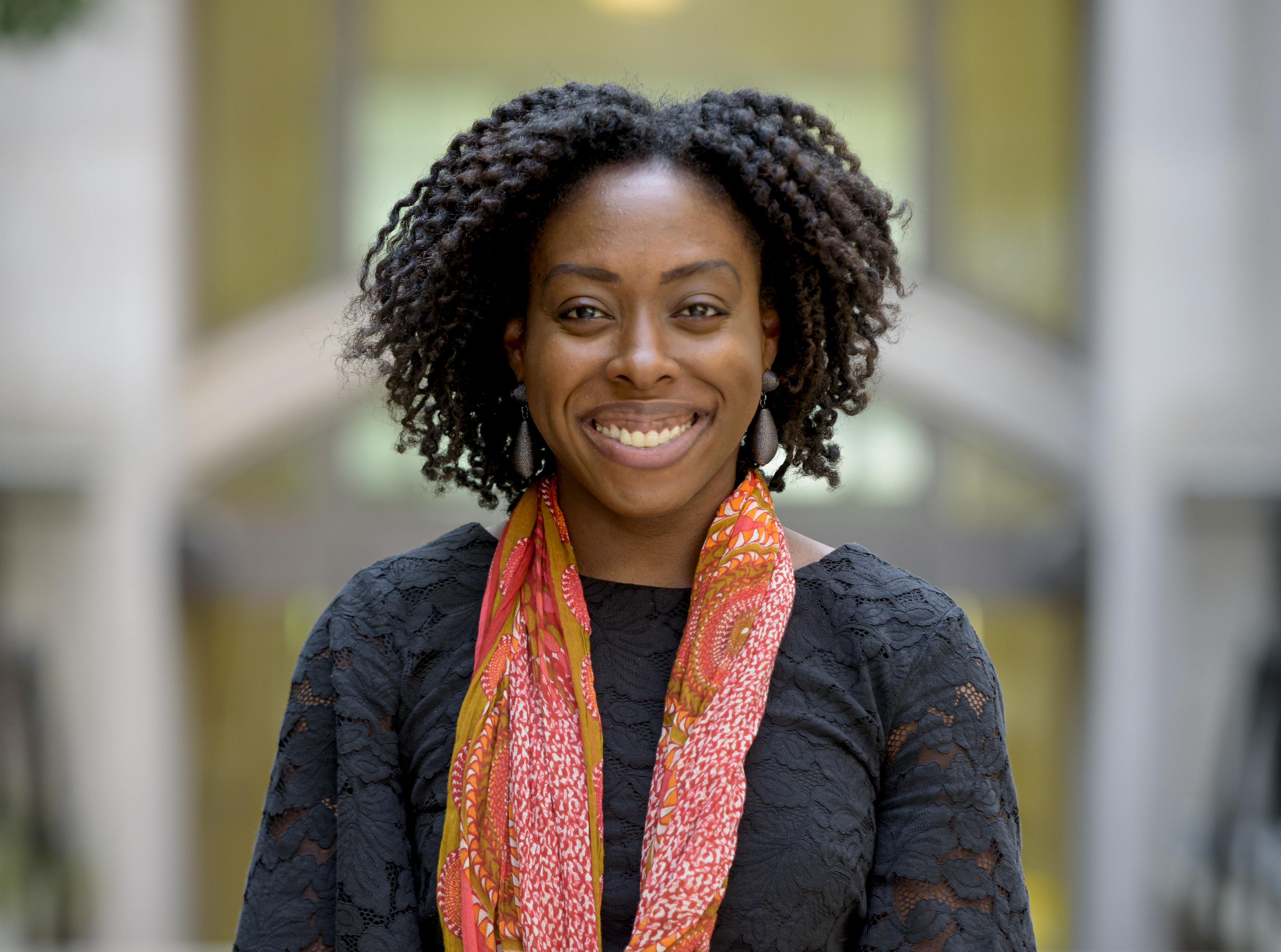 Robin Brewer, a black woman smiling with shoulder length curly hair wearing a black lace floral shirt and a patterned orange scarf.