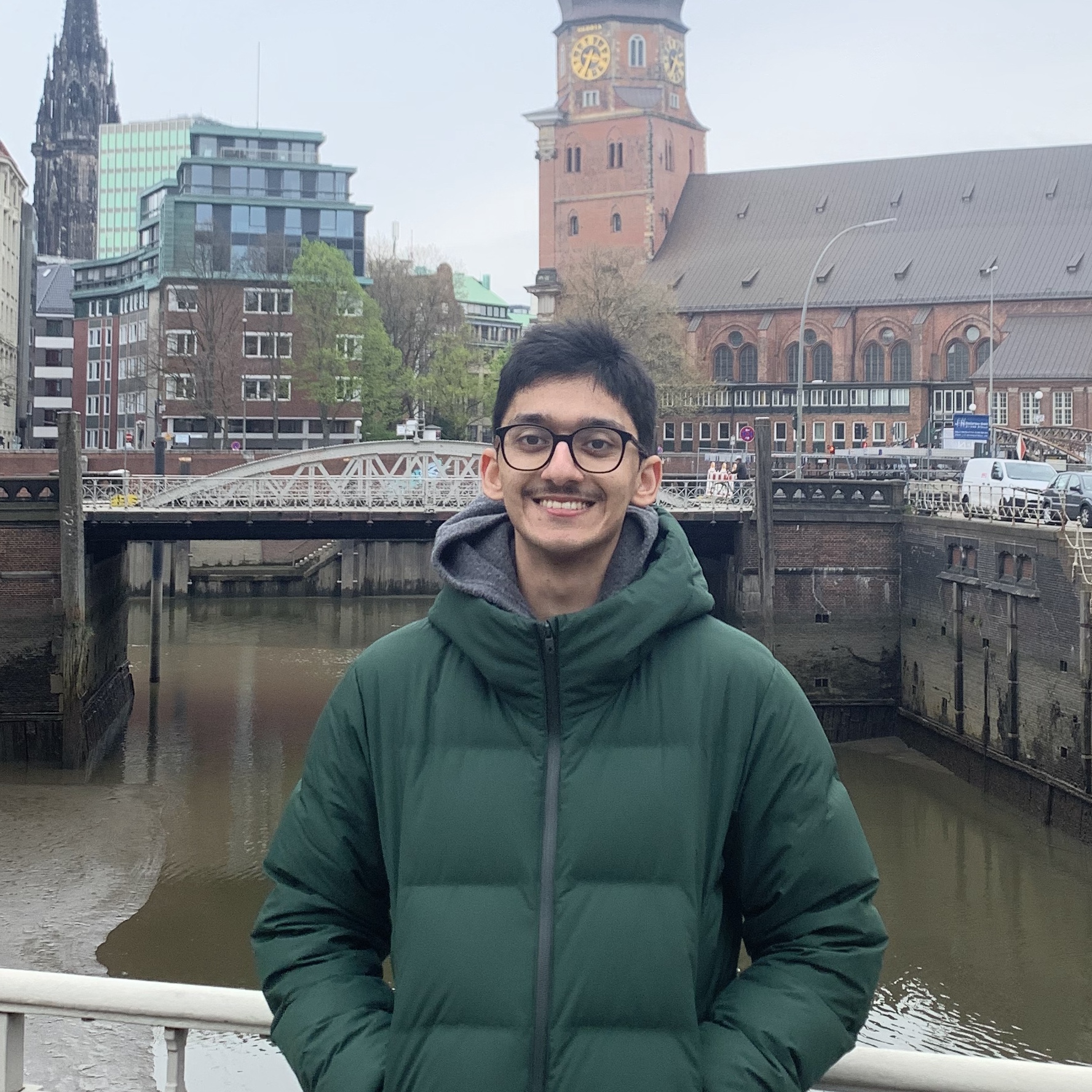 Portrait of Ryan Paul, a brown man with short dark hair and glasses. He is wearing a green winter jacket, smiling.