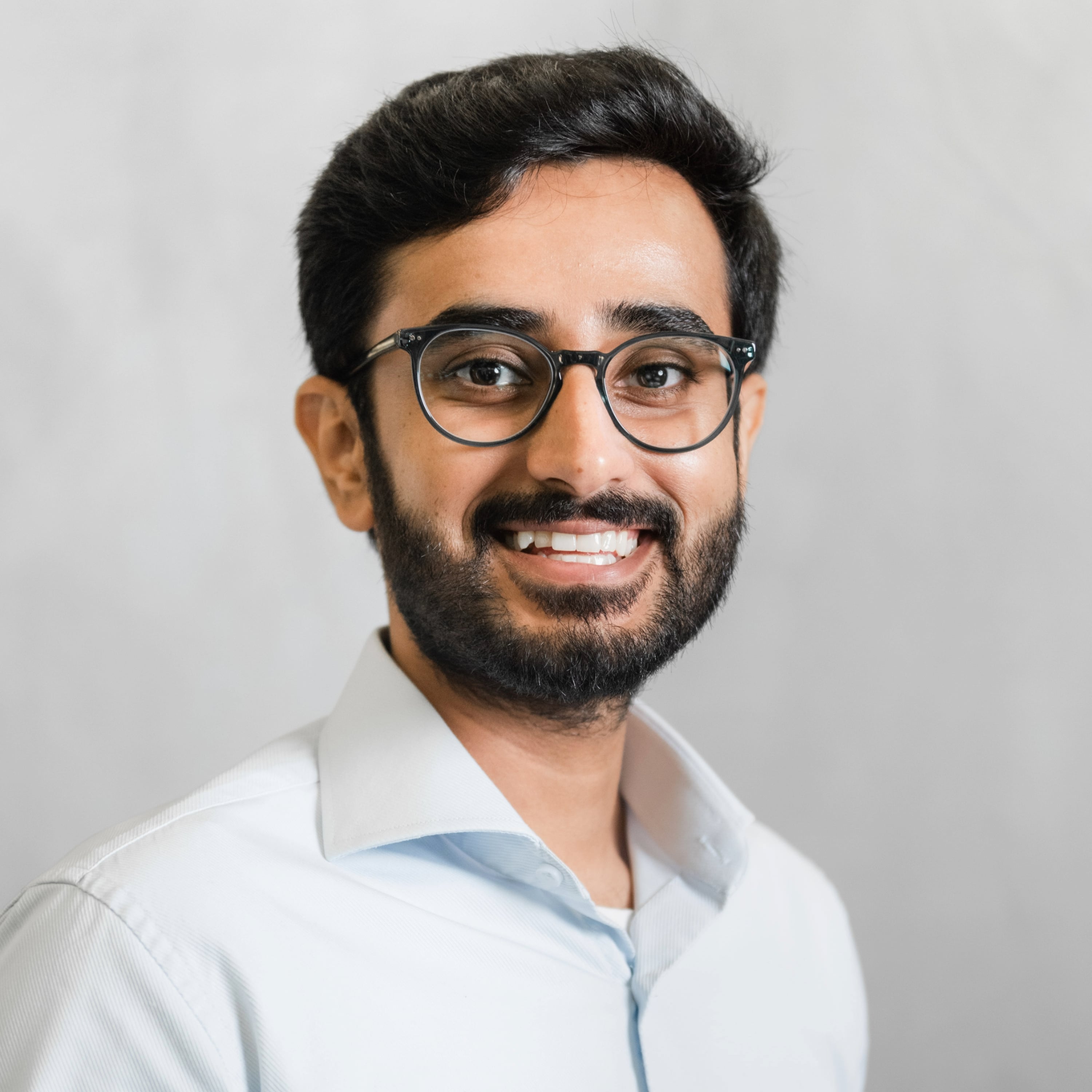 Portrait of Saad Hassan, a brown, black-haired male with a beard, wearing a light blue dress shirt and peacock green glasses, smiling.