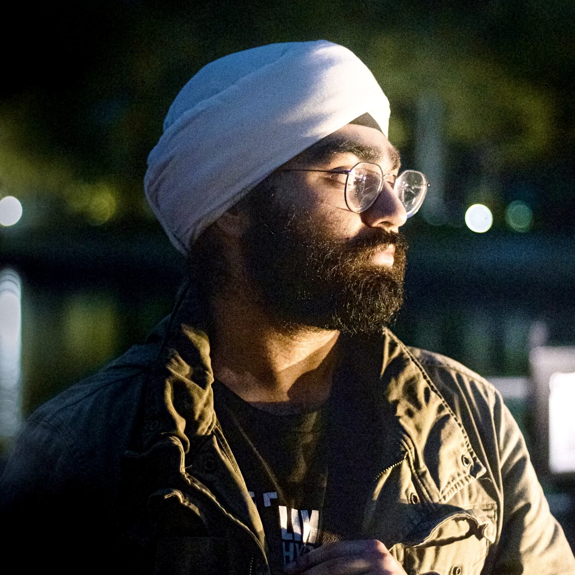 Portrait of Suraj Goraya, a brown man with a beard and glasses, wearing a white turban. He is looking towards the right side of the camera and is dressed in a green jacket over a black t-shirt.