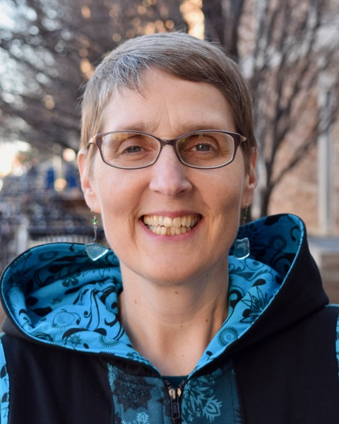 Portrait of a smiling woman with short grey hair and glasses, wearing a blue and black printed jacket, standing outside against a blurred background of trees.